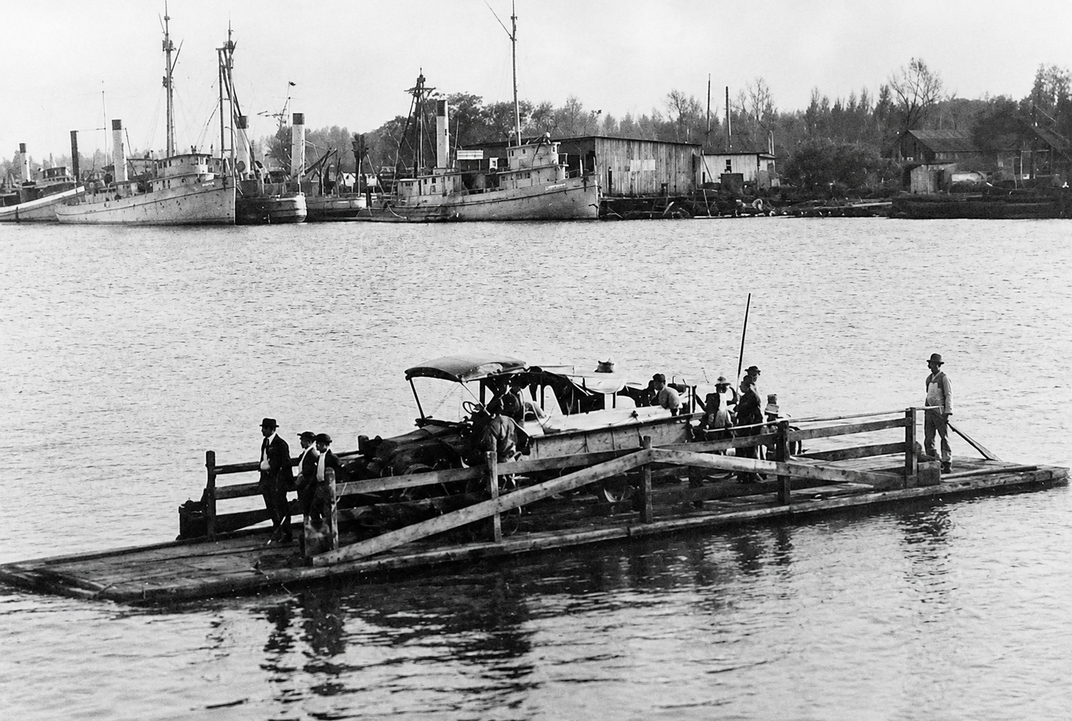 Historic ferry flat carrying passengers and an automobile across the Cape Fear River, with vessels and buildings along the Eagles Island waterfront